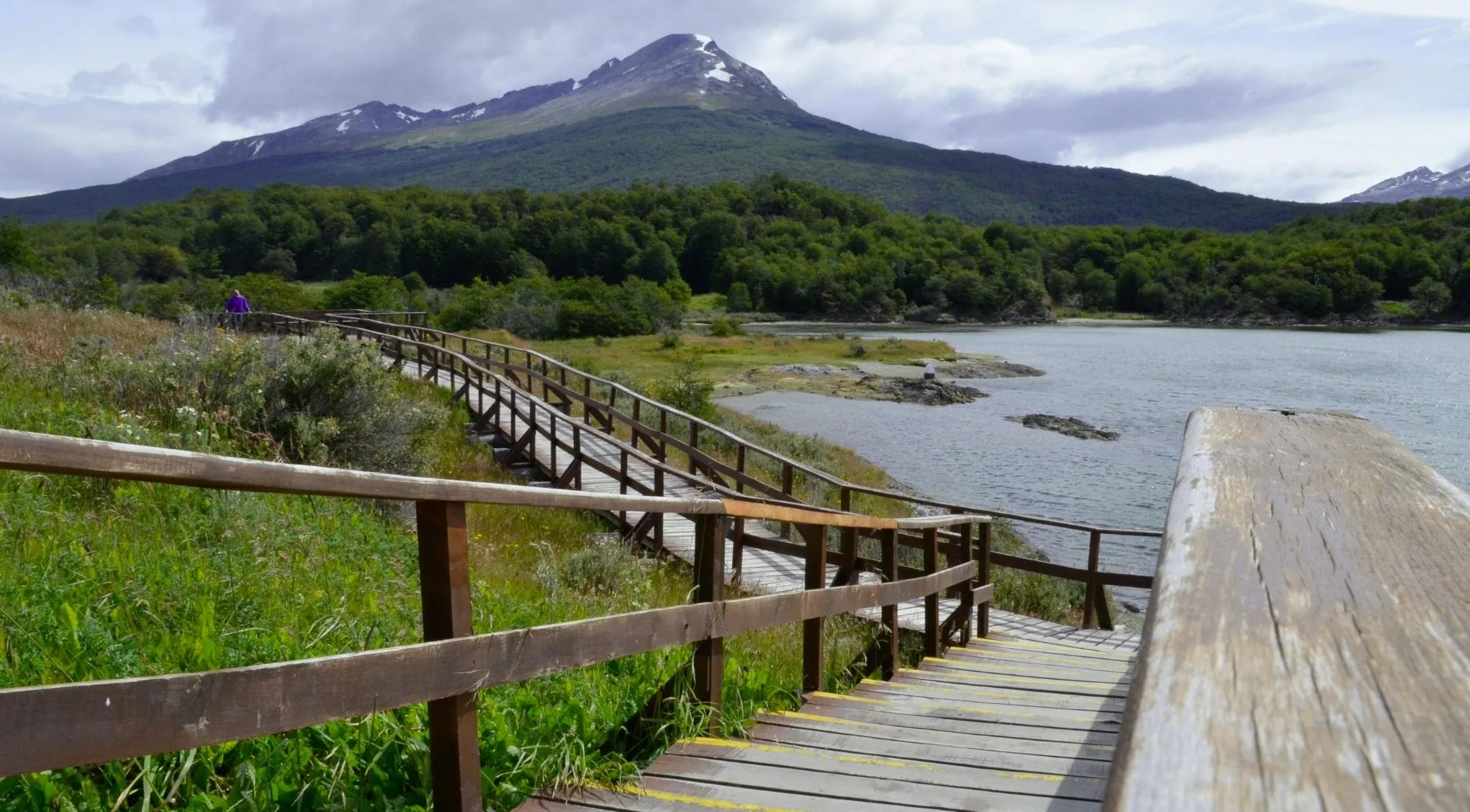 Wooden boardwalk and railings leading along a lake shore under a snow-streaked peak in Tierra del Fuego National Park