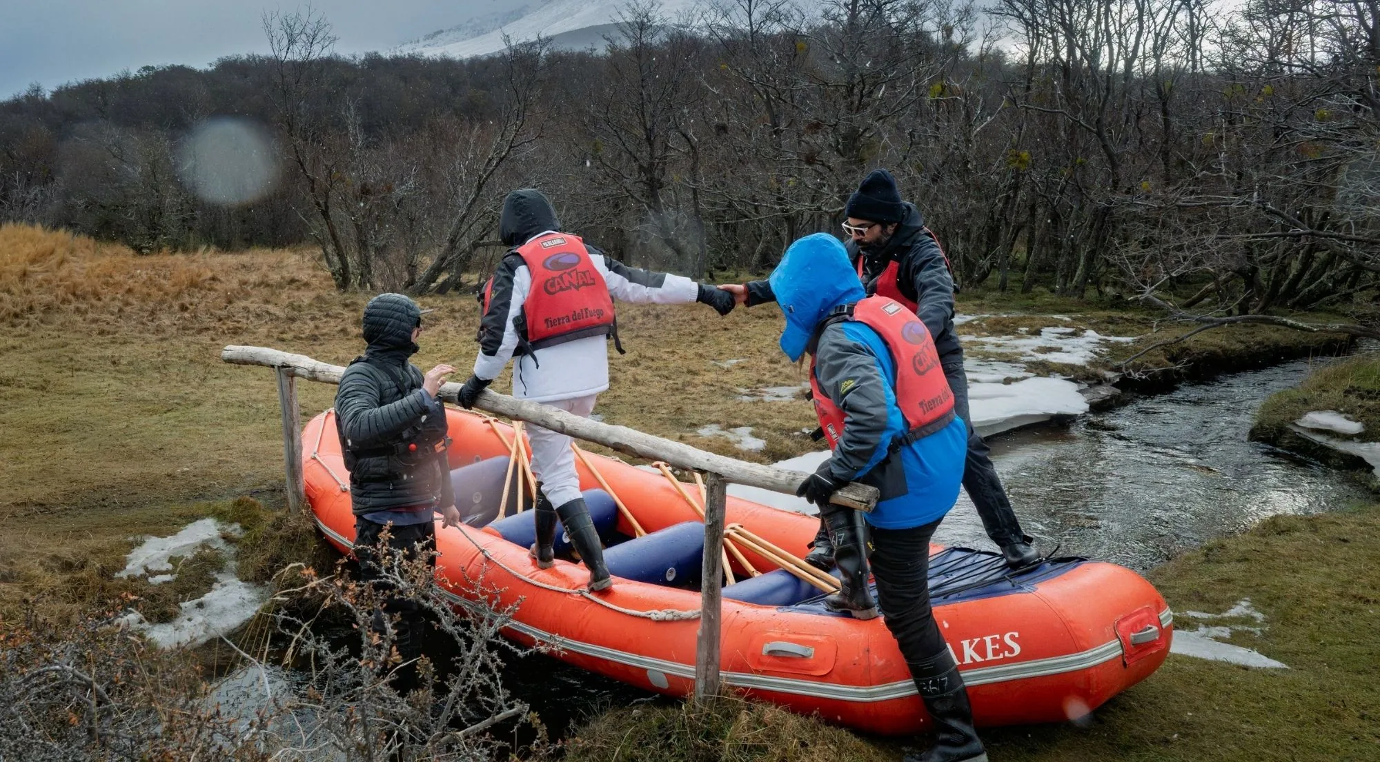 Four people in life jackets carrying an orange inflatable canoe across a stream through autumn grass