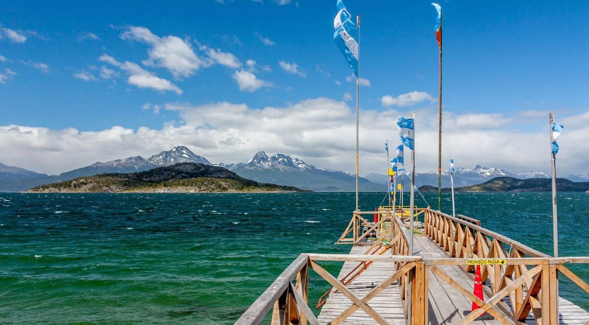 Wooden pier with Argentine flags reaching into turquoise water, with snow-capped mountains across the channel